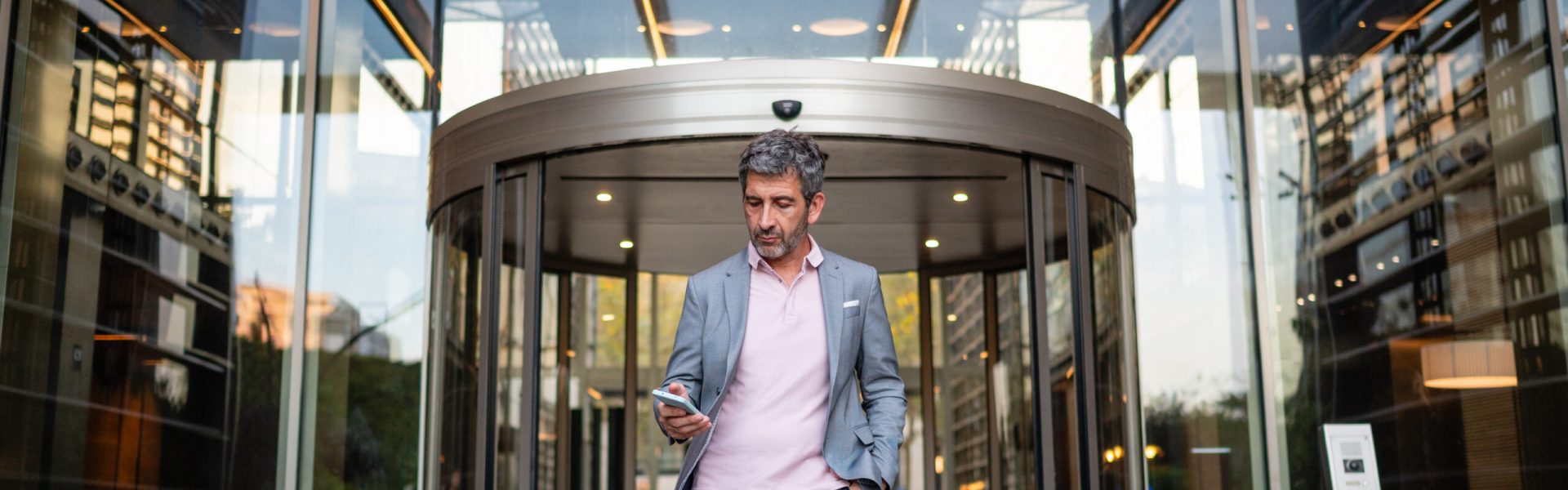 Professional businessman exiting a contemporary office building while checking his smartphone