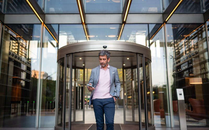 Professional businessman exiting a contemporary office building while checking his smartphone